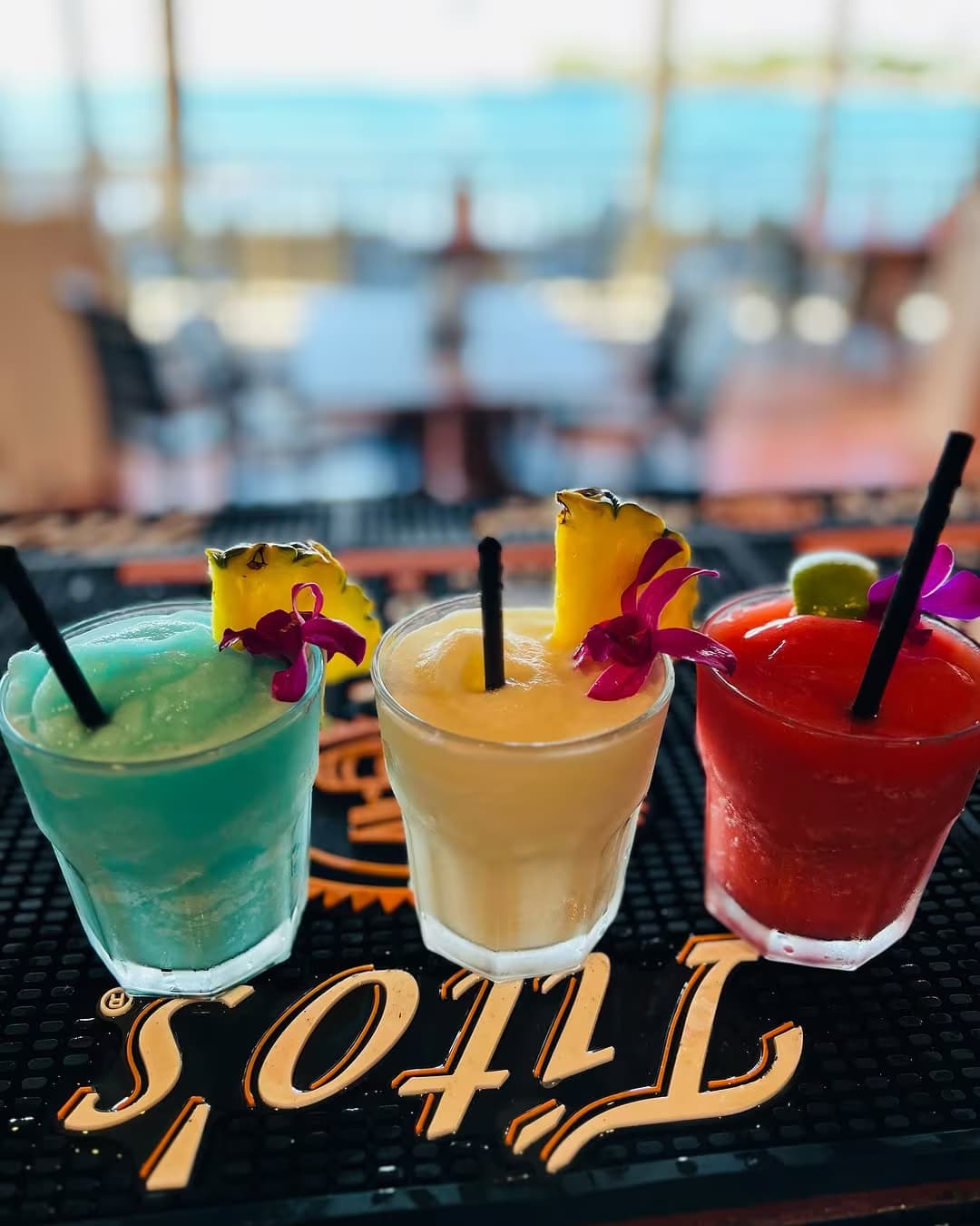 Three tropical frozen cocktails lined up on a bar mat with the Kona coast behind them