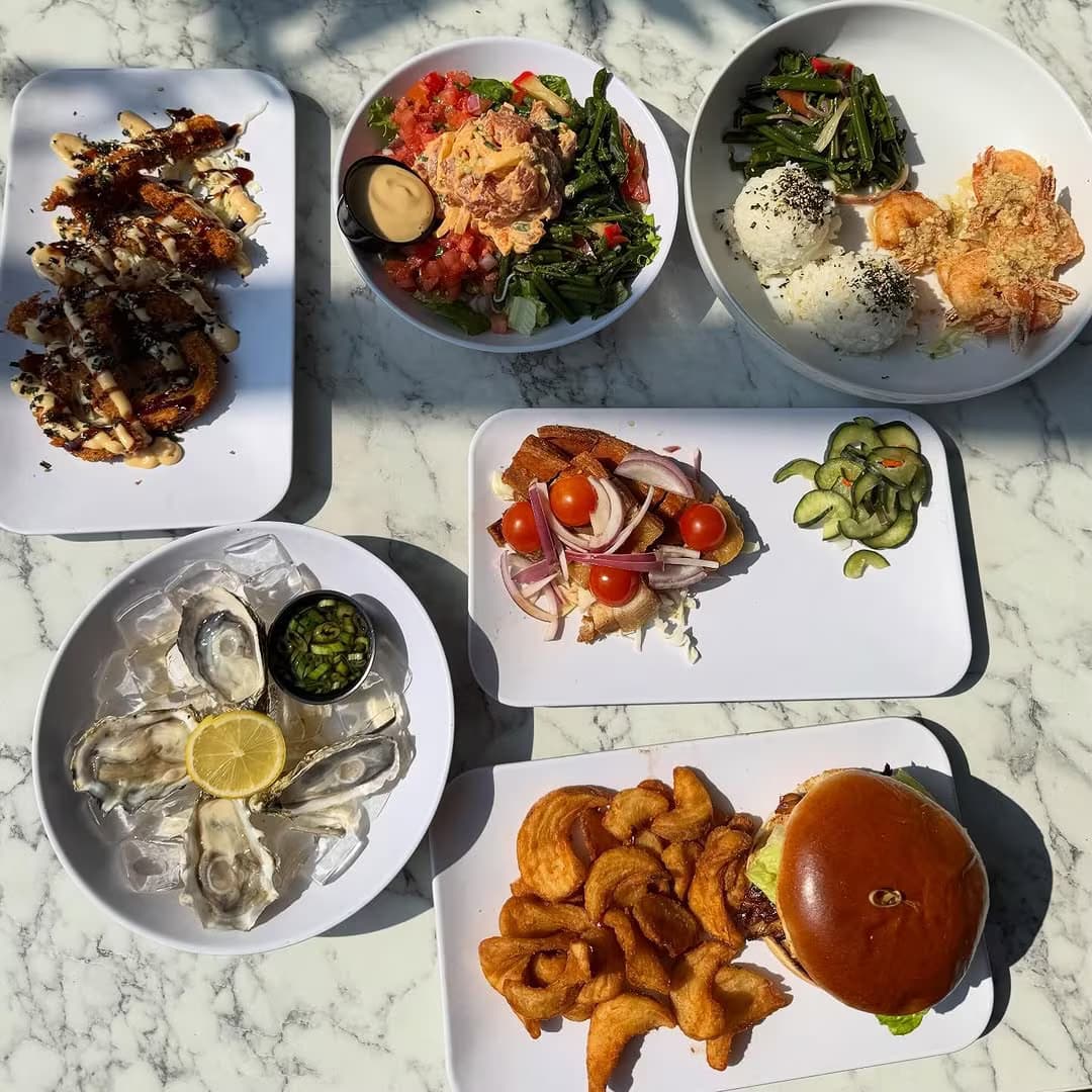 Overhead spread of burger, poke, oysters, salads, and tiradito on a marble table
